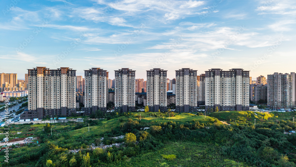 Fototapeta premium Modern high-rise residential buildings stand adjacent to a lush green urban landscape under a clear blue sky.