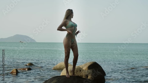 A young Caucasian female model stands elegantly on a stone by the beach posing in a stylish bikini as part of a professional fashion photoshoot. She gently touches swimwear and gazes toward skyline