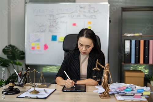 Focused professional writes on a tablet at her desk. She sits near symbolic legal objects. Whiteboard holds brainstorming notes and colorful stickers. Desk holds a scale, gavel, and small statue