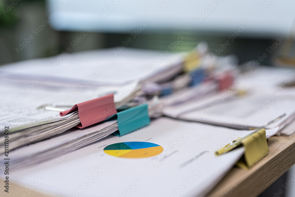Fototapeta premium Pile of papers rests on wooden desk surface. Colorful clip holds documents neatly together. Pie chart sits among papers, hinting at data analysis