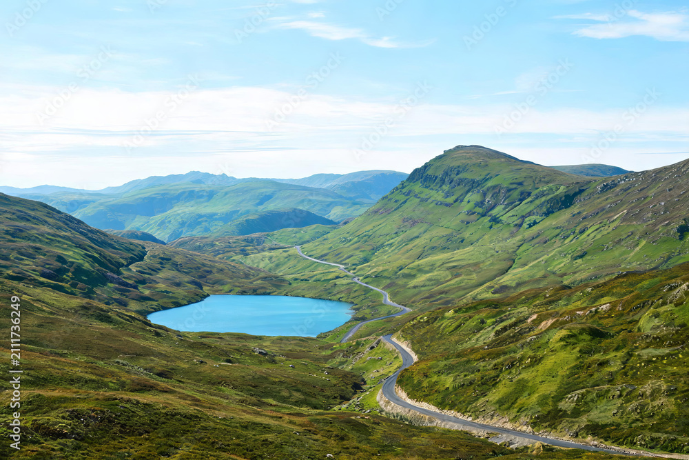Fototapeta premium A scenic highland landscape featuring a turquoise lake, winding mountain road, and rolling green hills under a clear blue sky.