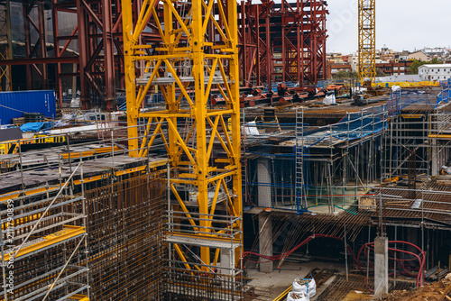 Yellow crane rises above red steel framing and rows of scaffolding across a wide building site. Large scale urban development and construction investment shaping skylines and property markets