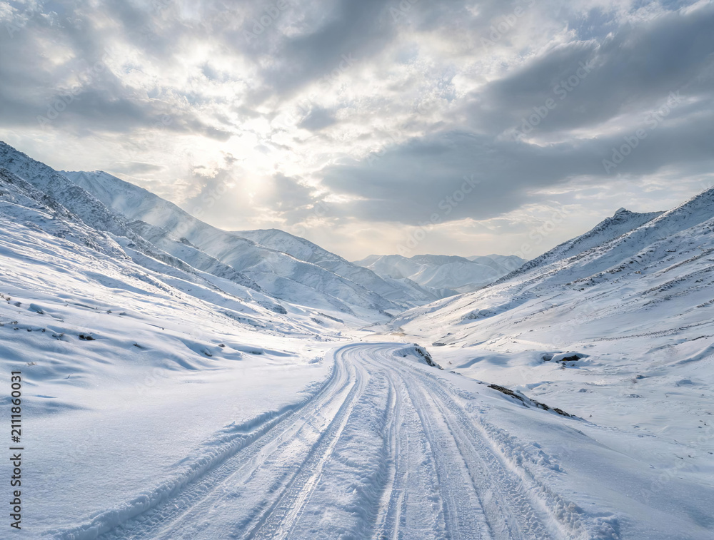 Fototapeta premium A winding snow track cuts through a vast snow-covered mountain valley under a partly cloudy winter sky with sunlight filtering through.