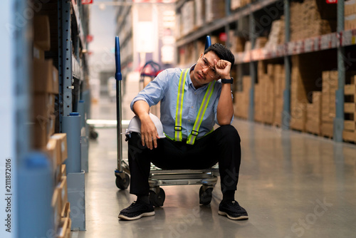 A male warehouse worker wearing a blue shirt is sitting down, appearing distressed or confused amidst the fatigue of warehouse work.