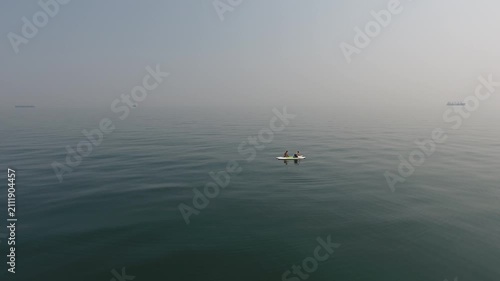 Drone view of two people on a stand up paddle board floating on calm open sea with distant cargo ships and soft haze creating a peaceful marine landscape