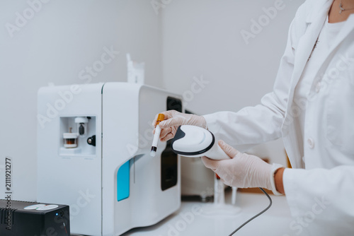 Lab Technician Scanning Blood Sample Vial With Handheld Scanner Next To Automated Clinical Analyzer