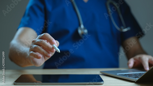 Male doctor wearing scrubs holds stylus pen over digital tablet screen working laptop computer inside dark medical office space night shift practitioner examining electronic health record data file.