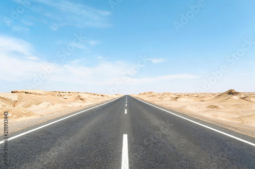 A straight empty asphalt highway stretches far into the horizon, crossing an arid desert under a clear blue sky.