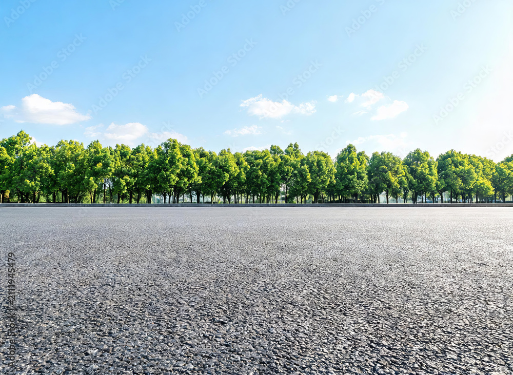 Fototapeta premium A wide smooth asphalt road stretches forward, bordered by a row of lush green trees under a clear blue sky with scattered white clouds, creating a serene outdoor landscape.
