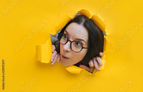 A portrait of a sweet young woman wearing glasses, looking through a hole in yellow paper. A surprised look. Feminine curiosity and gossip. An intellectual.