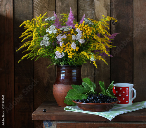 Wildflower bouquet with solidago and yarrow in a ceramic jug sits beside blackcurrants on a wooden table