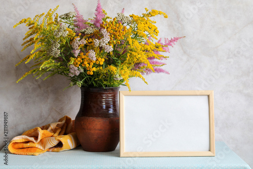 An empty mockup frame and a bouquet of flowers on the table: yarrow and Astilbe, goldenrod and tansy. Summer still life, cottage interior concept.