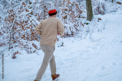 Фотография Young man in beanie and sweater shivers running in snowy forest, frosty morning,