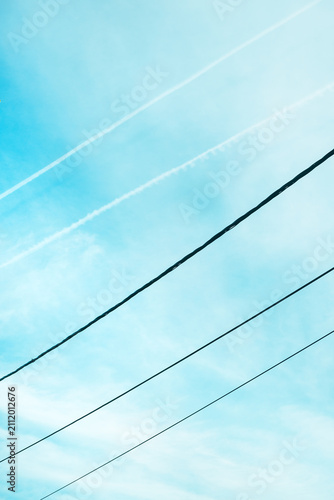 Minimal composition of a bright blue sky crossed by diagonal power lines and faint airplane contrails