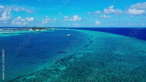 A serene aerial view of the Maldives, showcasing the tranquil tropical ocean and sky, with boats and a reef visible from a high-altitude perspective. The scene is bathed in sunlight, evocat.