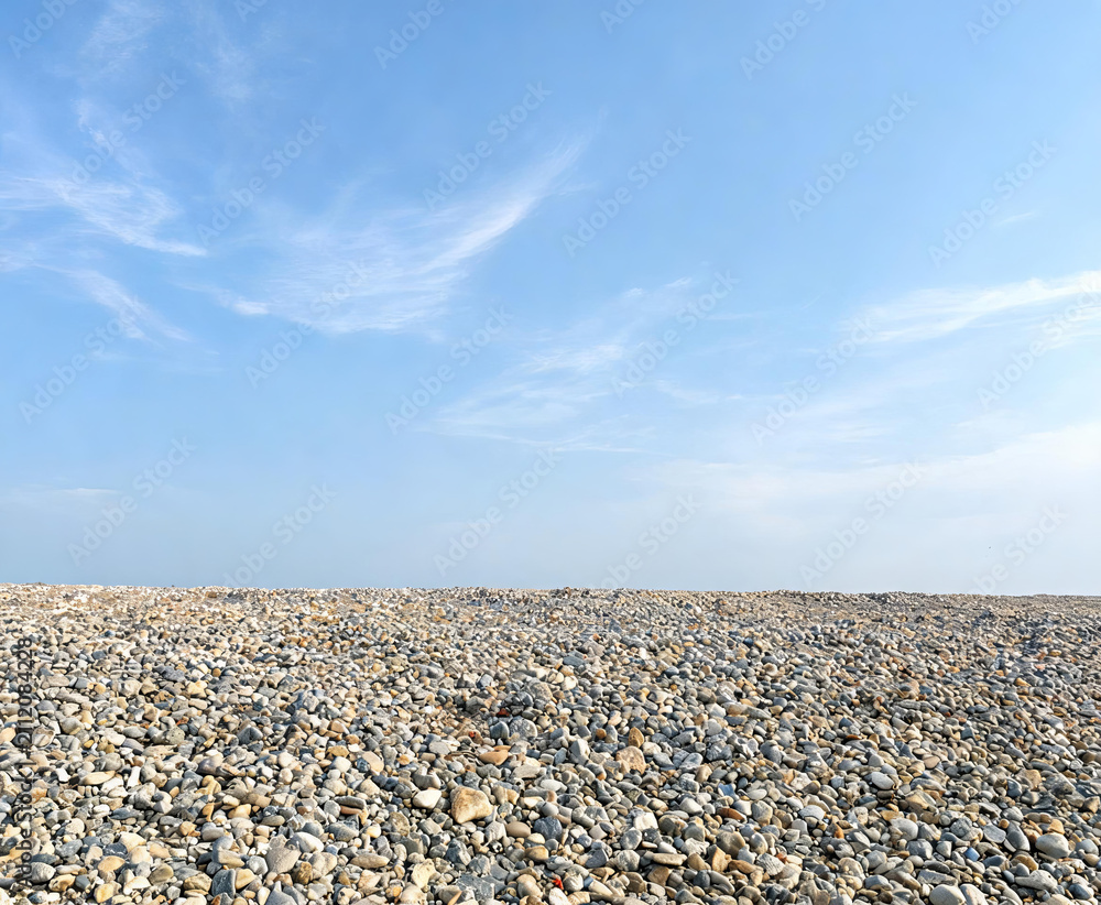 Obraz premium A vast expanse of smooth pebbles stretches to the horizon beneath a clear blue sky with wispy clouds.