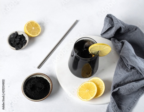 Activated charcoal lemon drink sits in a clear glass with ice and fresh lemon slices on a white table under soft natural sunlight