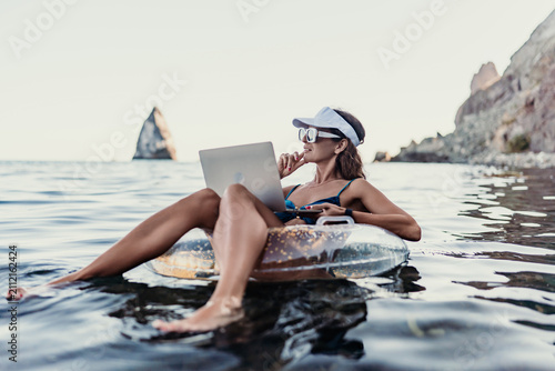 Business woman using laptop computer while floating on inflatable ring in calm ocean water near coastal cliffs