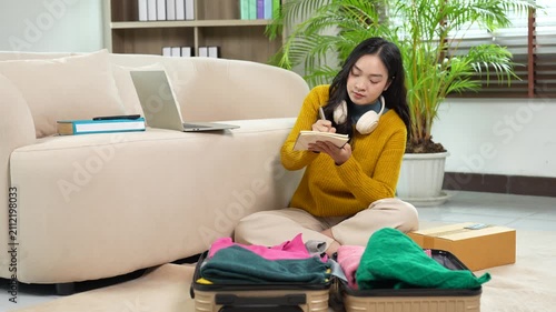 Young Asian woman planning her trip and packing her suitcase for winter vacation travel, writing a checklist of items to bring on her holiday journey in the living room