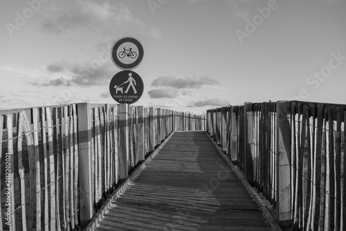 Black and white wooden boardwalk path leading through coastal dunes toward Labenne-Océan in the Landes, France, with regulatory signs. Monochrome landscape.