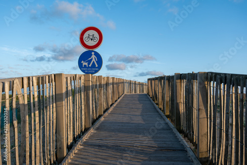 Wooden boardwalk path leading through coastal dunes toward Labenne-Océan in the Landes, France, with regulatory signs for bicycles and dogs on leash. Sunset lighting.