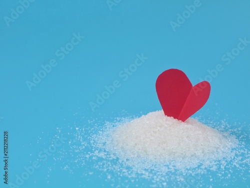 Photography Red paper heart standing in a pile of white granulated sugar on blue background,