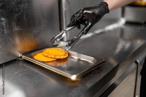 Konstfotografi A worker wearing a black glove uses tongs to pick up golden, crispy hash browns