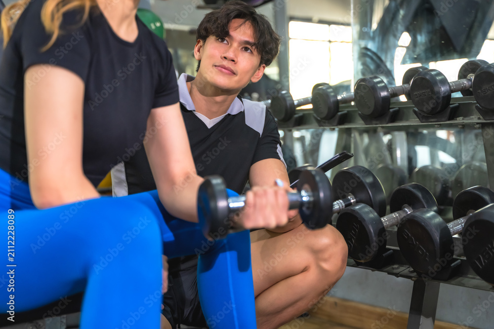 Fototapeta premium Handsome Asian male personal trainer helping and instructing a fit sportswoman during workout with dumbbells exercise equipment in modern gym