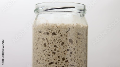Active sourdough starter with air bubbles fermenting in glass jar on white background.