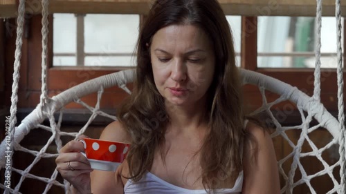 A thoughtful woman holds a red polka dot cup while looking down sadly. She sits inside a cozy macrame swing chair on a veranda.