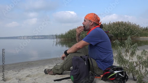Traveler resting on a quiet lakeshore while eating a sandwich during a cycling adventure. Peaceful wide shot with calm water, reeds, backpack, bicycle helmet, and relaxing outdoor atmosphere