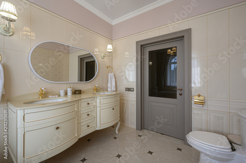 classic bathroom features a cream double‑sink vanity with curved legs, an oval mirror, gold fixtures, and beige tiles with dark star accents. wall sconces and towel rings add refined detail