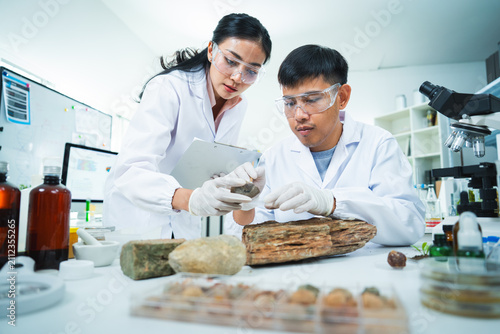 Scientist laboratory research specimen microscope safety goggle wood sample under careful study by Asian adult team in white coat with glove clipboard chemical bottle focused expression