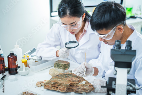 Scientist laboratory rock microscope geology research safety goggle student mineral sample examination during earth science study, focused teamwork in bright classroom workspace