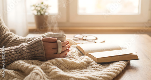 Hands wrapped in a cozy sweater holding a steaming mug beside an open book and reading glasses