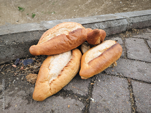 Pile of fresh bread loaves discarded on a paved city sidewalk next to a curb, with a blurred urban street background