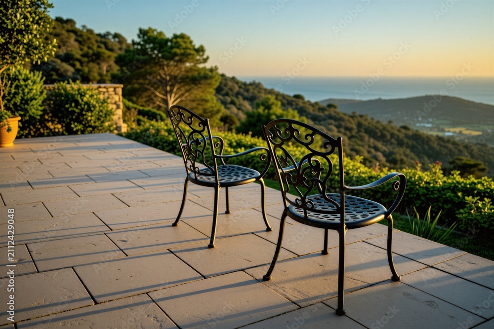Obraz premium Two metal chairs sit on stone terrace at sunset. Furniture faces scenic ocean view over green hills. Outdoor space provides calm atmosphere for relaxation on luxury resort patio. Empty seats wait for