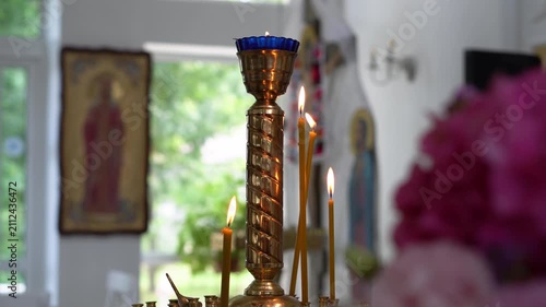 A close-up shot of thin yellow beeswax candles and a traditional brass oil lamp (vigil lamp) burning brightly inside an Orthodox Christian church. Orthodox icons and blurred flowers are visible in the