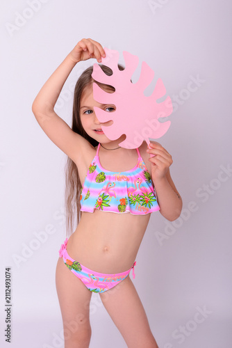 Photography Playful little girl in colorful swimwear hiding behind pink paper monstera leaf,