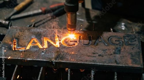 A plasma cutter precisely cuts the word 'LANGUAGE' into a metal sheet, creating brilliant sparks and molten material. Industrial metal fabrication technology in action.