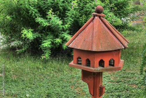 Hexagonal nesting box with multibple entrances, wooden material painted with brick red color, placed on wooden pole in public garden, summer cloudy sunshine. 