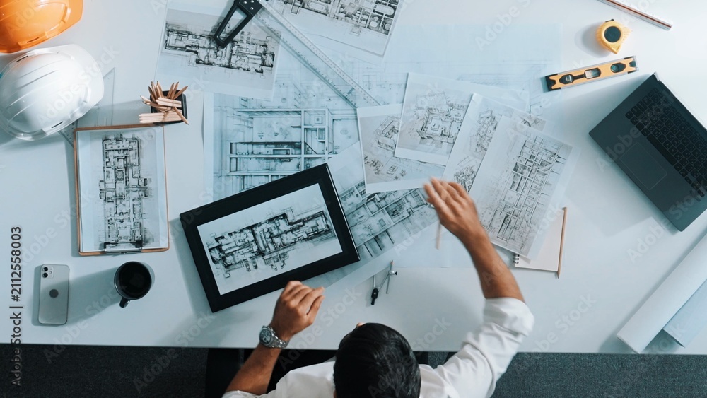 Obraz premium Top view of skilled engineer selecting material for house design while sitting at meeting table with tablet, laptop, safety helmet and equipment. Smart architect looking at project plan. Alimentation.
