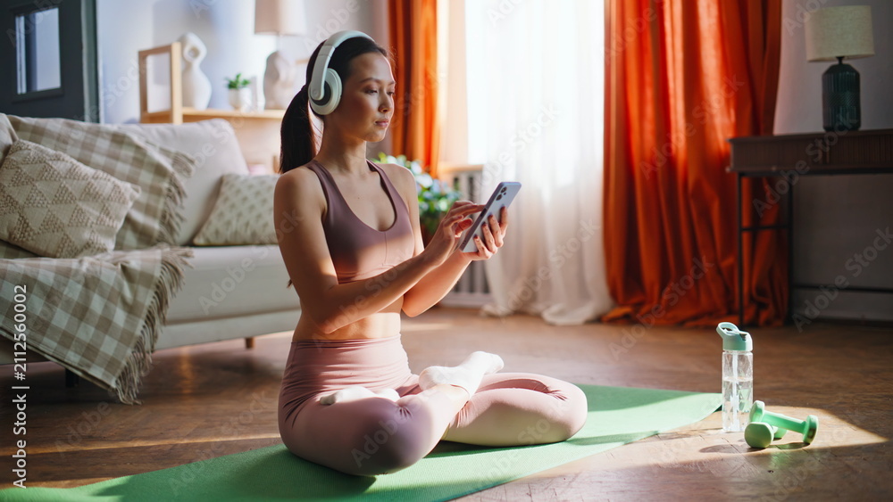 Fototapeta premium Sporty asian girl browsing phone before yoga meditation. Lotus pose home mat.