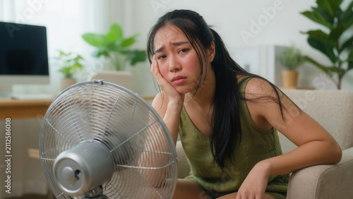 Fatigued Asian woman in hot home with fan, suffering from summer heat and seeking cooling relief