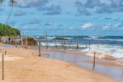 Traditional Sri Lankan stilt fishing posts along golden sandy beach and turquoise ocean waves in Ahangama. Wooden stilt fishing poles in the sea at Ahangama Beach, Sri Lanka. 