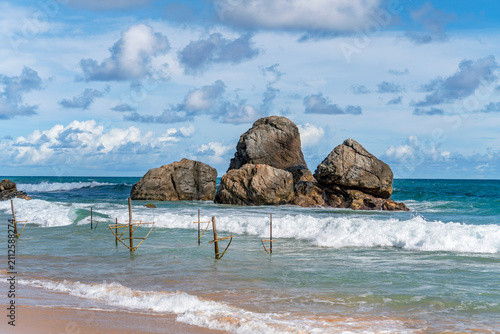 Traditional Sri Lankan stilt fishing posts along golden sandy beach and turquoise ocean waves in Ahangama. Wooden stilt fishing poles in the sea at Ahangama Beach, Sri Lanka. 
