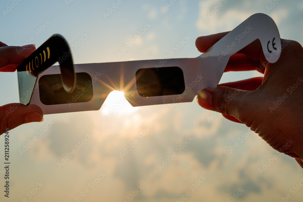 Obraz premium Hand holding certified solar eclipse viewing glasses against the bright sun in a vineyard landscape at sunset. The protective eclipse glasses are silhouetted by warm sunlight.