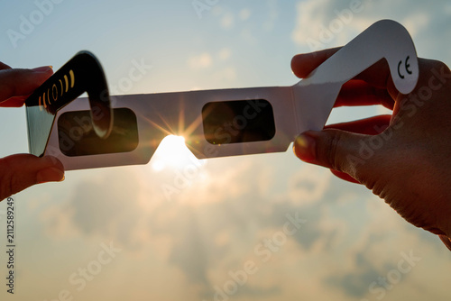 Obraz na plátně Hand holding certified solar eclipse viewing glasses against the bright sun in a vineyard landscape at sunset