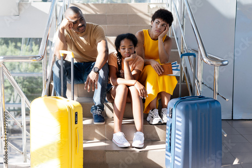 Tired African American family of three sitting on airport stairs, looking bored and exhausted while waiting near luggage suitcases, suffering flight delay