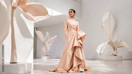 Elegant fashion portrait of a woman in a flowing peach gown surrounded by oversized abstract flowers in a modern studio. Ideal for editorials, luxury campaigns, bridal branding and creative advertisin